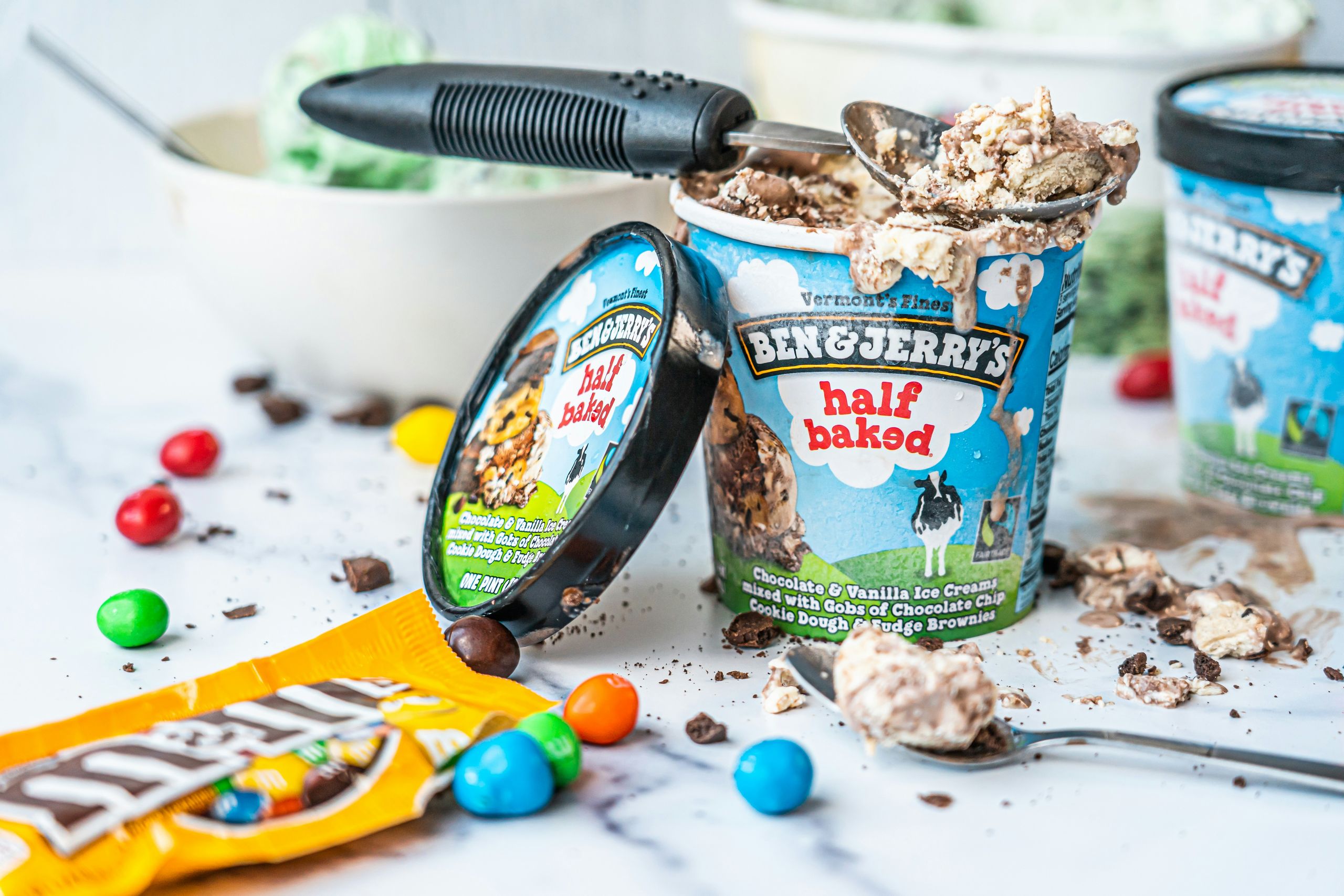 An open tub of Ben & Jerry's Half Baked ice cream with a scoop, chocolate pieces and sweets scattered around it