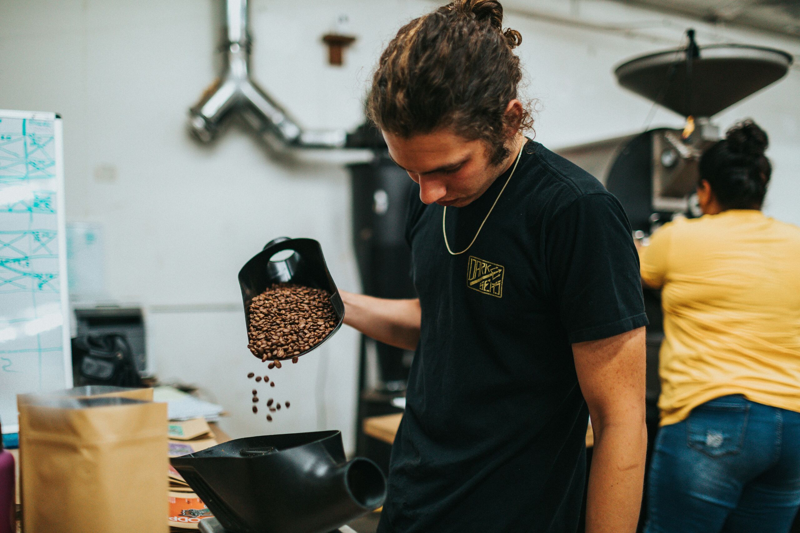 A man pouring freshly roasted coffee beans from a scoop in a roastery, a colleague working behind him