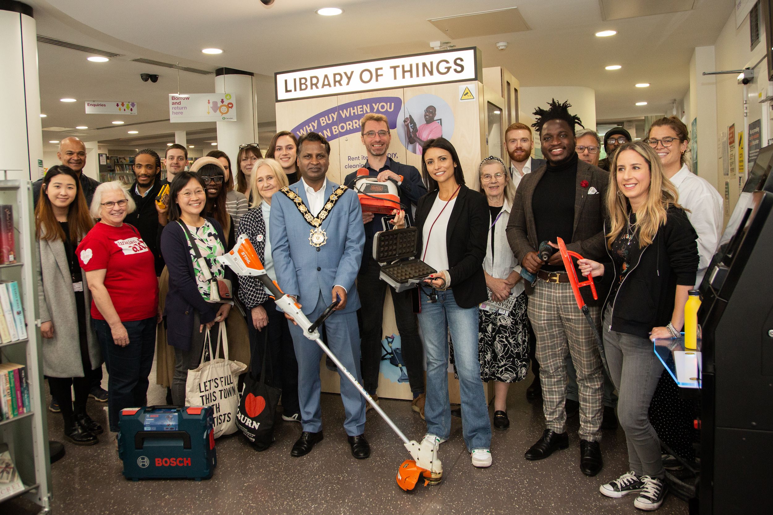 A crowd gathered in a library, a mayor in his chain of office at the centre, holding borrowed tools
