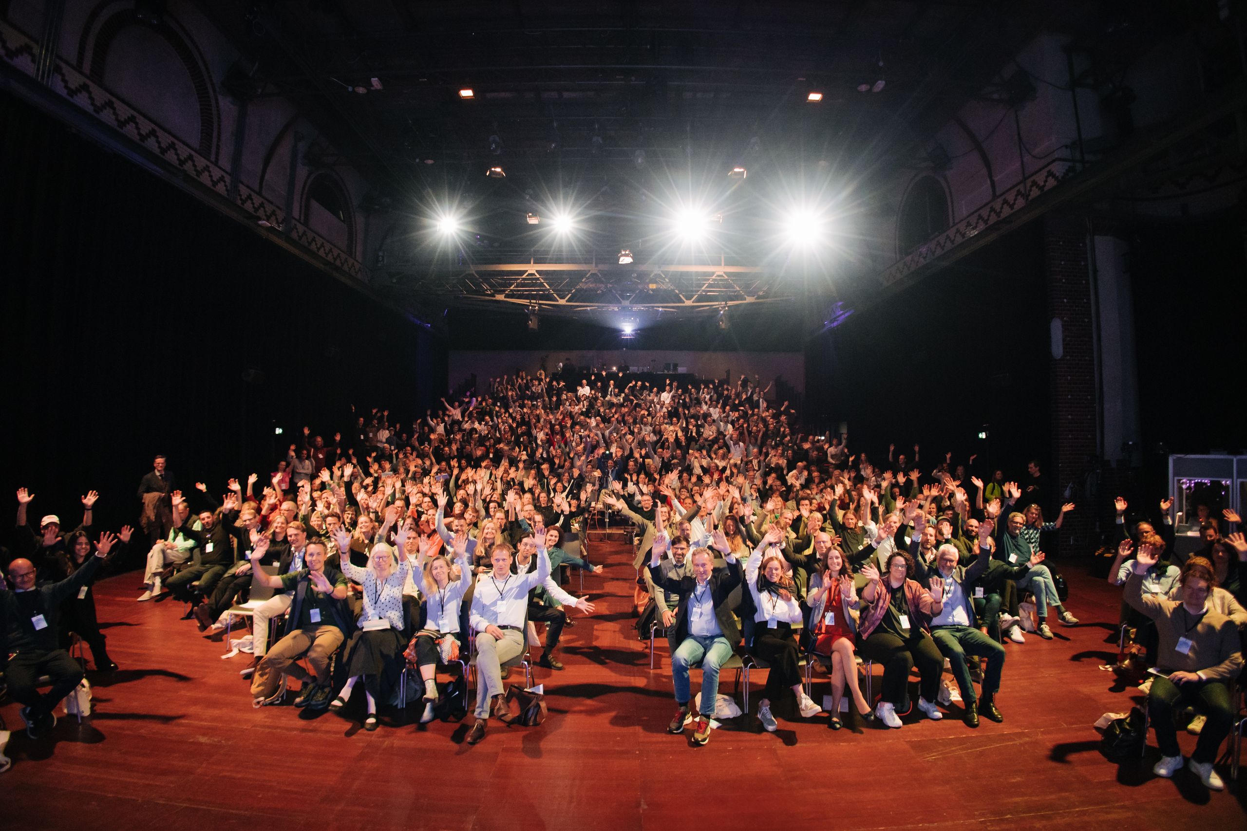 A packed conference hall of people waving towards the camera under stage lights